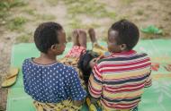 Two women laugh with one another, their backs turned towards the camera. They sit on a blanket, practicing their vocational skill of hair dressing; Photo credit: Serrah Galos