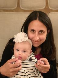 A woman with long dark hair looks up and smiles at the camera, with her baby daughter on her lap