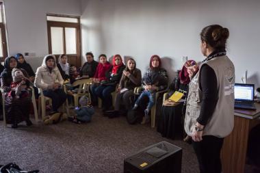 A classroom of women sits listening in a circle around their trainer