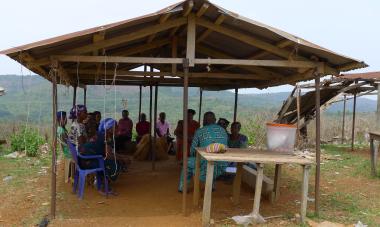 A group of people having a discussion underneath a shelter in Nigeria with a table of snacks. 