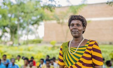 Caritas standing in a patterned shirt off to the right, looking proudly into the camera. Behind her is a class she trains at Women for Women International in Rwanda.