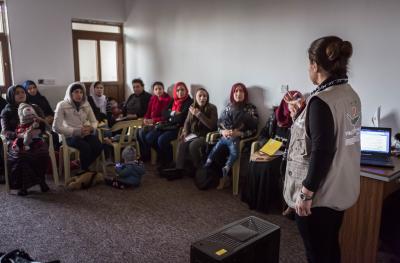 A classroom of women sits listening in a circle around their trainer